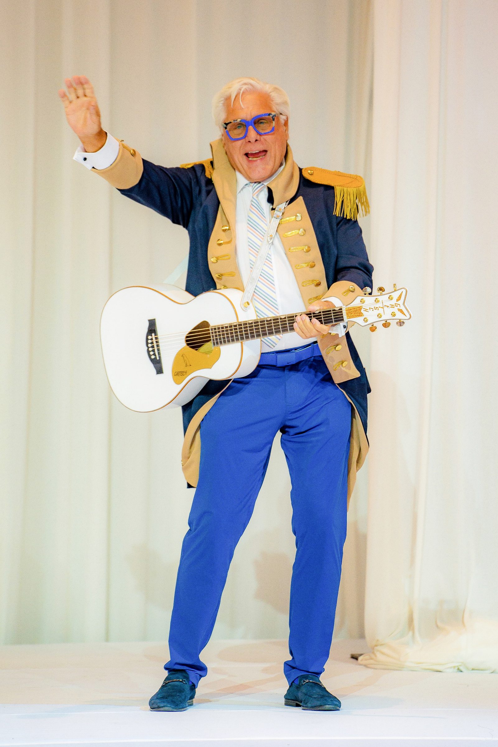 Robert Dempster waving in a navy and gold jacket, blue trousers and glasses, holding a white guitar